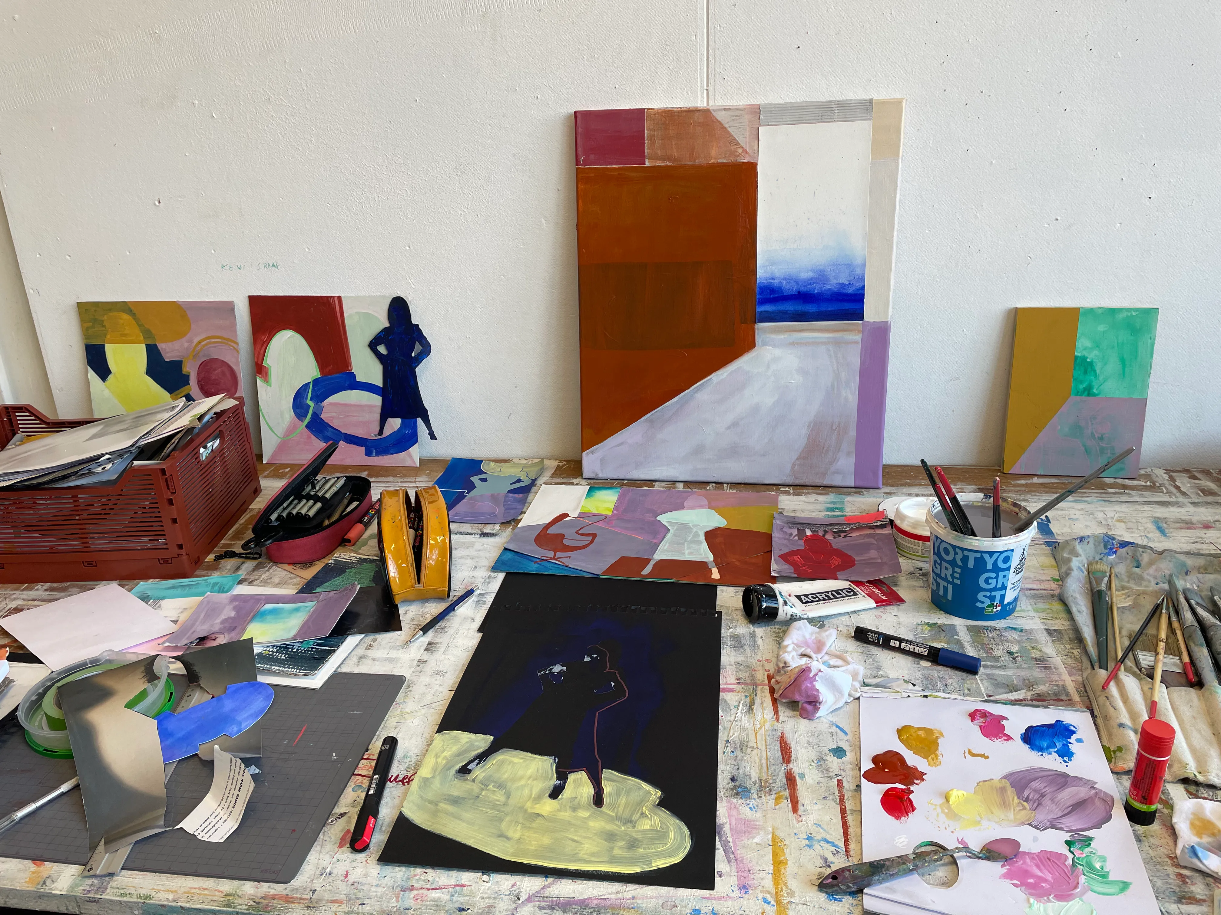 A worktable in the studio, spattered with paint and covered in studies on paper, brushes and palettes; four finished canvases lean against the wall behind it.